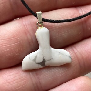 May include: White and gray whale tail pendant on a black cord. The pendant is made of a marbled stone and has a silver-colored bail. The whale tail design is a popular nautical theme.