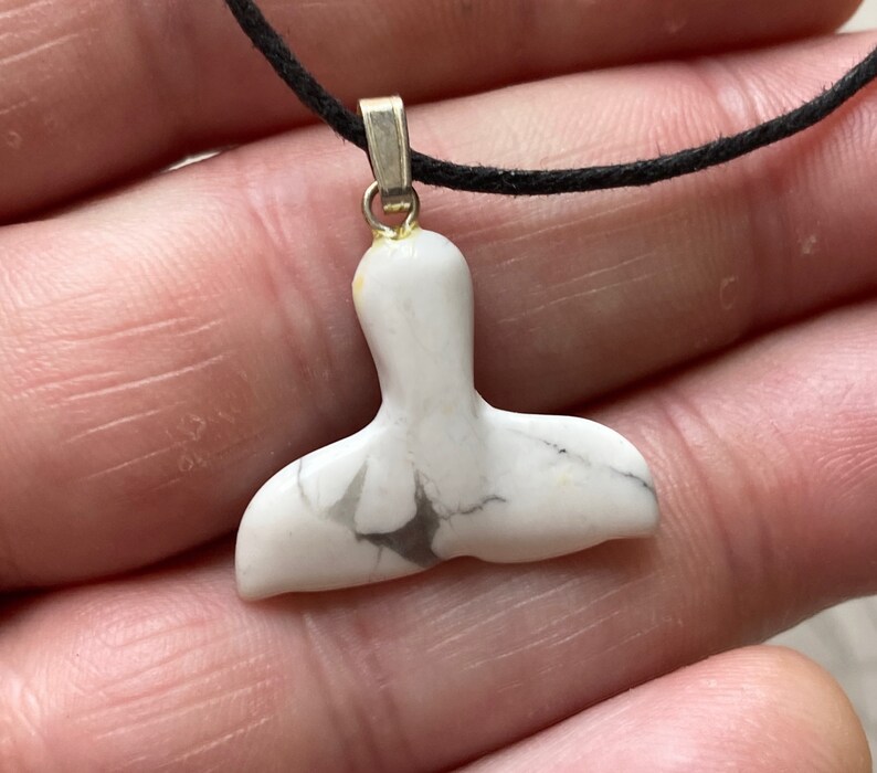 May include: White and gray whale tail pendant on a black cord. The pendant is made of a marbled stone and has a silver-colored bail. The whale tail design is a popular nautical theme.