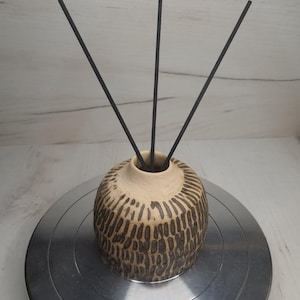 Może przedstawiać: Beżowy ceramiczny dyfuzor olejków eterycznych z teksturowanym, ciemnobrązowym wzorem. Trzy czarne patyczki dyfuzora są włożone w otwór. Dyfuzor stoi na srebrnej metalowej powierzchni.