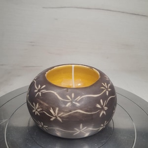 Op de afbeelding: Een ronde, donkerbruine keramische kandelaar met een gele binnenkant. De buitenkant heeft een herhalend wit bloemenpatroon. Een witte kaars staat in het midden, wat een warme sfeer creëert. Een decoratief item.
