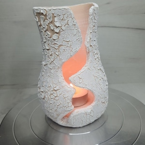 May include: A white ceramic candle holder with a textured, cracked surface. The holder has a curved, organic shape with a large, open design. A lit candle inside casts a warm glow, creating a cozy ambiance.
