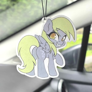 May include: A hanging car air freshener featuring a cartoon illustration of a grey pony with a light yellow mane and tail, large amber eyes, and small wings. The pony has a playful expression and is depicted in a standing pose with a white border.