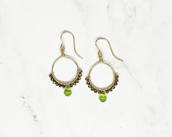 May include: Gold hoop earrings with a small green gemstone dangling from the bottom. The hoops are decorated with small gold beads.