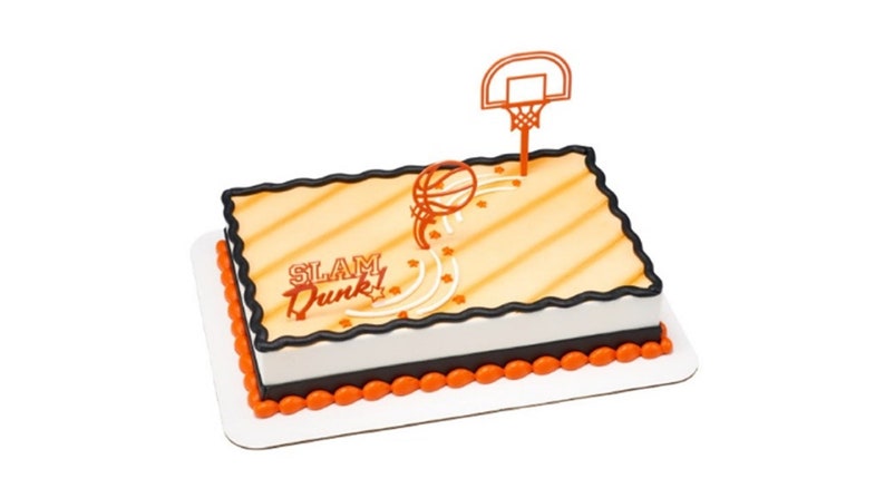 Peut inclure: Un g&acirc;teau carr&eacute; d&eacute;cor&eacute; d'un motif de terrain de basket-ball, d'un ballon de basket et d'un panier de basket. Le g&acirc;teau est glac&eacute; en orange et blanc avec une bordure noire et les mots "SLAM Dunk!" en orange.