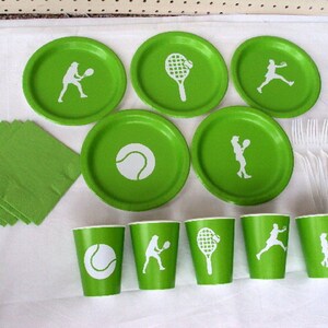 May include: Green and white paper plates and cups with tennis-themed designs. The plates feature a tennis ball, a tennis racket, and a tennis player. The cups feature a tennis ball, a tennis racket, and a tennis player. There are also green napkins and white plastic forks.