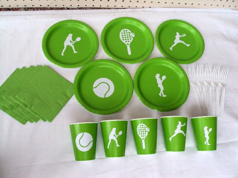 May include: Green and white paper plates and cups with tennis-themed designs. The plates feature a tennis ball, a tennis racket, and a tennis player. The cups feature a tennis ball, a tennis racket, and a tennis player. There are also green napkins and white plastic forks.
