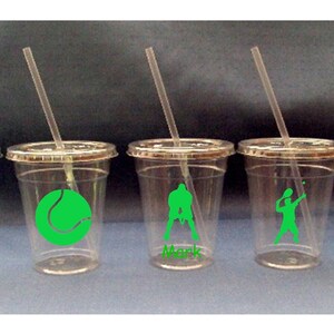 May include: Five clear plastic cups with green tennis-themed designs and names on the bottom. The cups have lids and straws. The designs include a tennis racket, a tennis ball, a tennis player serving, a tennis player hitting a volley, and two tennis players playing doubles.