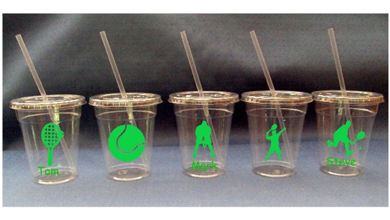 May include: Five clear plastic cups with green tennis-themed designs and names on the bottom. The cups have lids and straws. The designs include a tennis racket, a tennis ball, a tennis player serving, a tennis player hitting a volley, and two tennis players playing doubles.