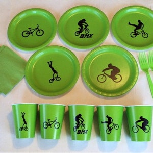 May include: Green paper plates and cups with black silhouettes of BMX riders and bikes. The plates have the word "BMX" printed on them. There are also green napkins and forks.