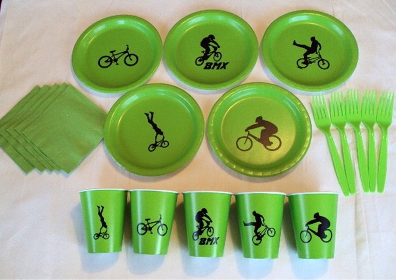 May include: Green paper plates and cups with black silhouettes of BMX riders and bikes. The plates have the word "BMX" printed on them. There are also green napkins and forks.