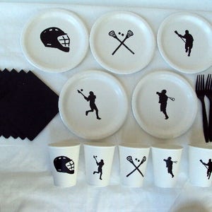 May include: A white lacrosse-themed party set featuring plates, cups, napkins, and forks. The plates and cups have black lacrosse graphics, including helmets, players, and crossed sticks. Black napkins and forks complete the set.
