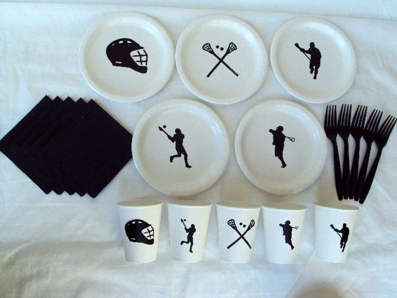 May include: A white lacrosse-themed party set featuring plates, cups, napkins, and forks. The plates and cups have black lacrosse graphics, including helmets, players, and crossed sticks. Black napkins and forks complete the set.