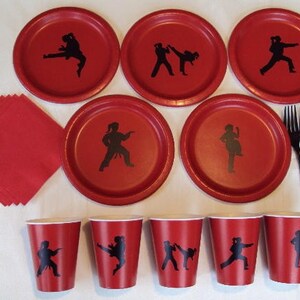 May include: Red and black paper plates and cups with a silhouette of a person doing a karate kick. There are also black plastic forks.