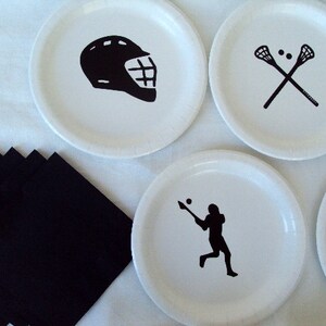 May include: White paper plates with black lacrosse-themed graphics, including a helmet, crossed sticks, and a player in action. Black napkins are arranged on the left. Ideal for a sports-themed party or event.