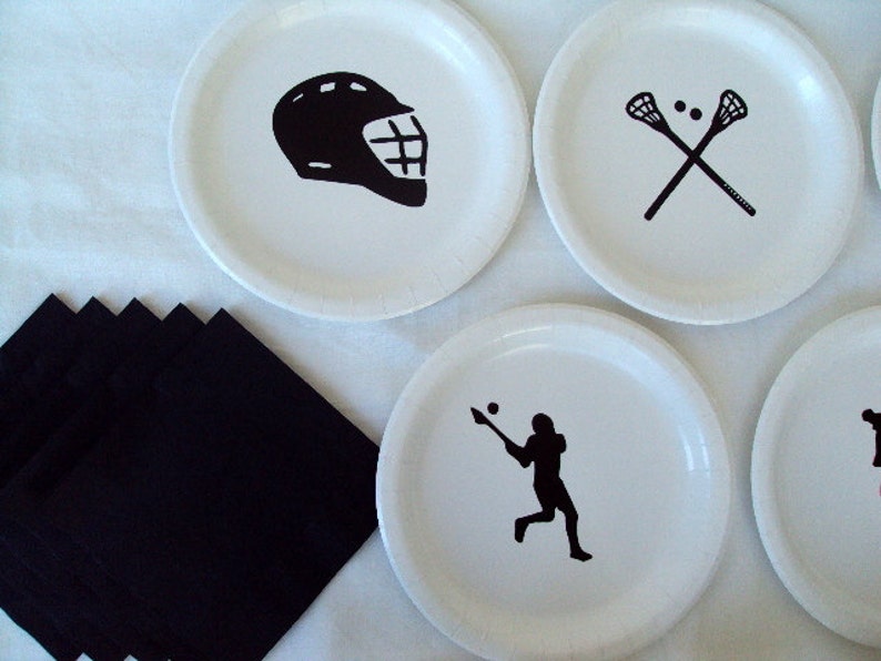 May include: White paper plates with black lacrosse-themed graphics, including a helmet, crossed sticks, and a player in action. Black napkins are arranged on the left. Ideal for a sports-themed party or event.