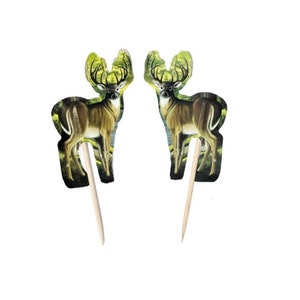 May include: Two brown deer cupcake toppers with green foliage backgrounds. The deer are standing on wooden skewers.