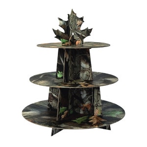 May include: Three-tiered cake stand with a camouflage pattern. The stand is made of cardboard and features a leaf-shaped topper.