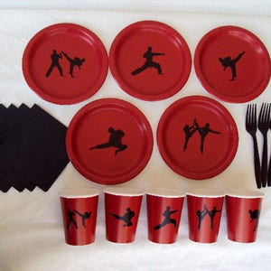 May include: Red paper plates with black silhouettes of martial arts figures. The plates are arranged in a circle with black napkins and black plastic forks in the center.