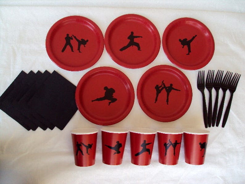 May include: Red paper plates with black silhouettes of martial arts figures. The plates are arranged in a circle with black napkins and black plastic forks in the center.