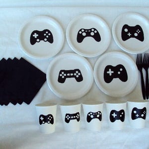 May include: A set of white paper plates and cups with black video game controller designs. The plates are 9 inches in diameter and the cups are 3.5 inches tall. There are also black napkins and black plastic forks.