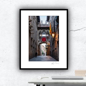 May include: Framed print of a narrow European street scene. A stone bridge with ornate arches and a red floral decoration spans the street. A Catalan flag hangs from a balcony. A person walks down the cobblestone street.