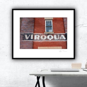 May include: A vintage brick building with a faded sign that reads "VIROQUA". The sign is painted in black and white letters on a red brick wall. There is a single window in the building, with a white frame and a view of the sky.
