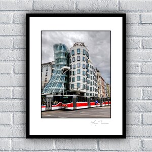 May include: Framed print of the Dancing House in Prague, Czech Republic. The modern building has a unique, deconstructivist design with a glass tower and a white stone facade. A red and white tram is in front of the building.