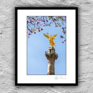 May include: A framed print showing a golden angel statue on a tall column against a blue sky. The angel wears a laurel wreath. The image is framed in black and includes a signature at the bottom.