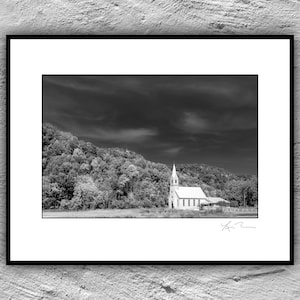 May include: A black and white photograph of a small white church with a steeple, surrounded by trees. The sky is cloudy and dramatic.