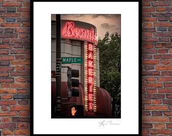 May include: A framed photograph of a neon sign for "Bennis Bakeries" at a street corner. The sign is red and the building is beige. A street sign reads "Maple Ave". A hand signal for "stop" is lit.