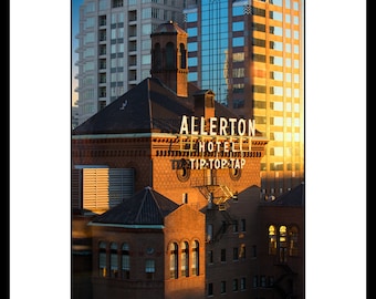 May include: A brick building with a sign that reads "Allerton Hotel Tip-Top Tap" in white letters. The building is illuminated by the setting sun, casting a warm glow on the brick facade. The building is surrounded by other buildings, including a modern glass building.