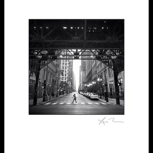 May include: A black and white photograph of a person walking across a city street under an elevated train track. The street is lined with tall buildings and cars are parked on the side of the street.