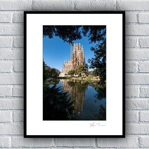 May include: A framed photograph of the Sagrada Familia basilica in Barcelona, Spain. The basilica is partially obscured by trees, and its reflection is visible in a pond in the foreground.