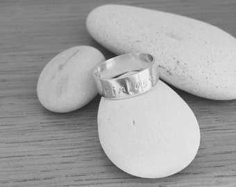 May include: A silver ring with the name "Finlay" engraved on it. The ring is resting on two smooth, white stones.