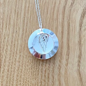 Puede incluir: Un collar con colgante de plata con un colgante circular con un diseño detallado de ala. El colgante cuelga de una delicada cadena de plata. El collar se muestra sobre una superficie de madera.