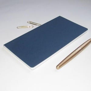 May include: A navy blue notebook with a gold pen and two paper clips. The notebook is rectangular and has a smooth, dark blue cover. The pen is metallic gold and cylindrical. The paper clips are silver.