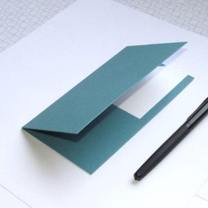 May include: A teal-colored, folded paper document holder with a white interior, next to a black pen. The holder is open, revealing the inside. The pen is sleek and modern, with a matte finish. The background is white with a subtle pattern.