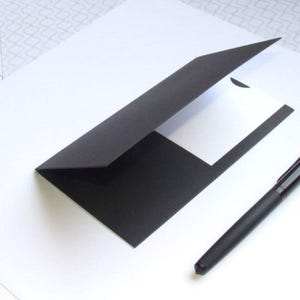 May include: A black presentation folder with a white insert and a black pen. The folder is open, revealing the white interior. The pen is lying next to the folder on a white surface.