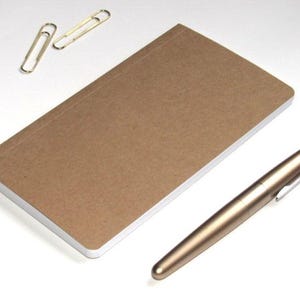 May include: A brown notebook with a white-edged cover, a gold pen, and two gold paper clips are arranged on a white surface. The pen is metallic and has a silver clip. The notebook is rectangular.