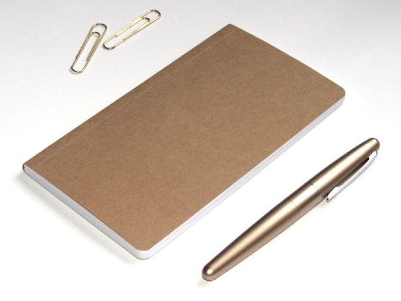 May include: A brown notebook with a white-edged cover, a gold pen, and two gold paper clips are arranged on a white surface. The pen is metallic and has a silver clip. The notebook is rectangular.