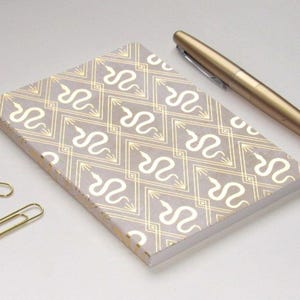 May include: A rectangular notebook with a light gray cover featuring a repeating gold foil pattern of snakes inside diamond shapes. A metallic gold fountain pen rests diagonally above the notebook, and two gold paper clips are positioned to the left.