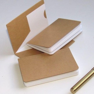 May include: A set of small, rectangular notebooks with brown kraft paper covers and white pages. One notebook is open, revealing a pocket inside the cover, while another is closed. A gold-toned metal pen and two silver paper clips are nearby.