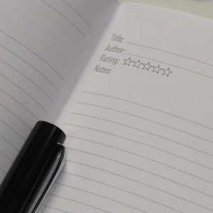 May include: An open notebook with lined pages and a black pen. The notebook has prompts for title, author, rating, and notes. The rating section includes five star outlines. The pen is black with a silver clip.
