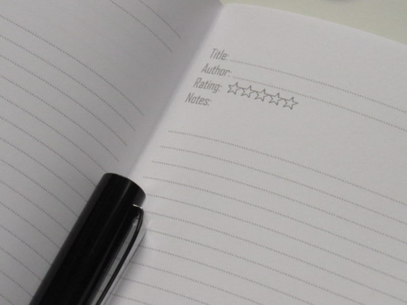 May include: An open notebook with lined pages and a black pen. The notebook has prompts for title, author, rating, and notes. The rating section includes five star outlines. The pen is black with a silver clip.