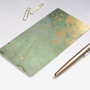 May include: A rectangular notepad with a green and gold watercolor design. A gold pen and two gold paper clips are next to the notepad. The notepad is approximately 8 inches long and 4 inches wide.