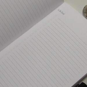 May include: Open notebook with lined pages and the text "To Be Read" in the upper left corner. A silver and gold coin is in the upper right corner. The notebook is open to a blank page.