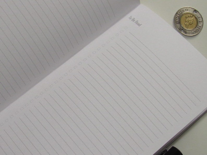 May include: Open notebook with lined pages and the text "To Be Read" in the upper left corner. A silver and gold coin is in the upper right corner. The notebook is open to a blank page.