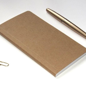 May include: A brown rectangular notebook with a gold pen and two gold paper clips on a white surface. The notebook is open, revealing white pages. The pen is metallic and cylindrical.