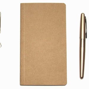 May include: A tan-colored notebook, a gold pen, and two gold paper clips are arranged on a white background. The notebook is rectangular and closed. The pen is metallic and has a clip. The paper clips are in the upper left.
