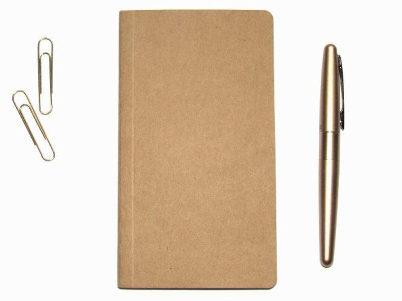 May include: A tan-colored notebook, a gold pen, and two gold paper clips are arranged on a white background. The notebook is rectangular and closed. The pen is metallic and has a clip. The paper clips are in the upper left.